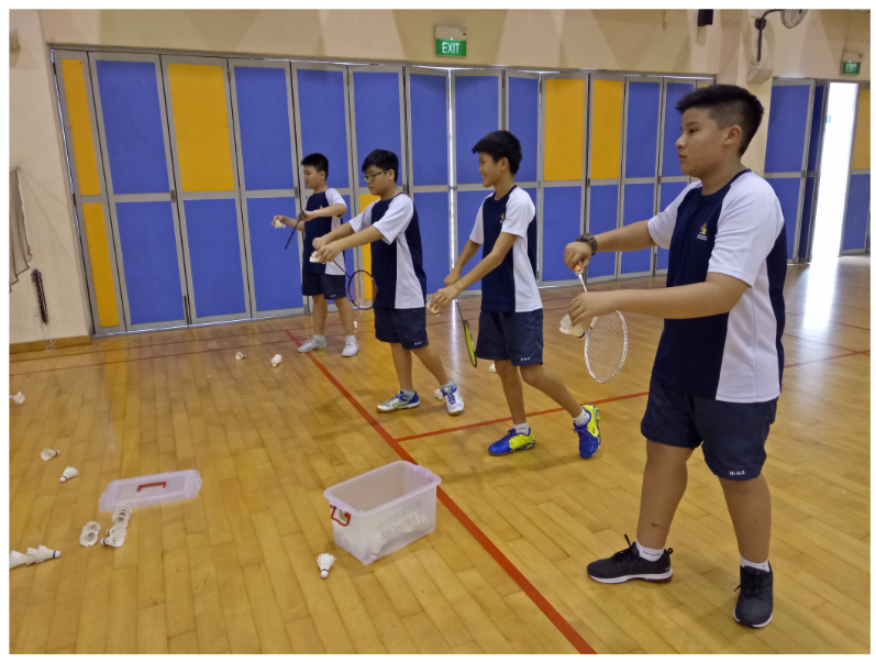 Four boys in school uniforms practice badminton with rackets and shuttlecocks on an indoor wooden court.